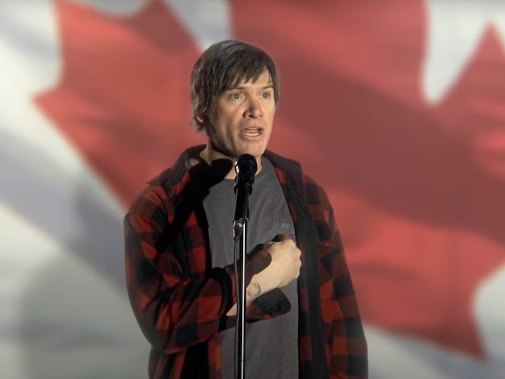 Jeff Douglas has shaggy dirty-blond hair and is wearing a red buffalo plaid shirt over a dark grey T-shirt. He stands at a microphone in front of a large projected image of the Canadian flag. He is mid-speech, one hand pointing to his chest.