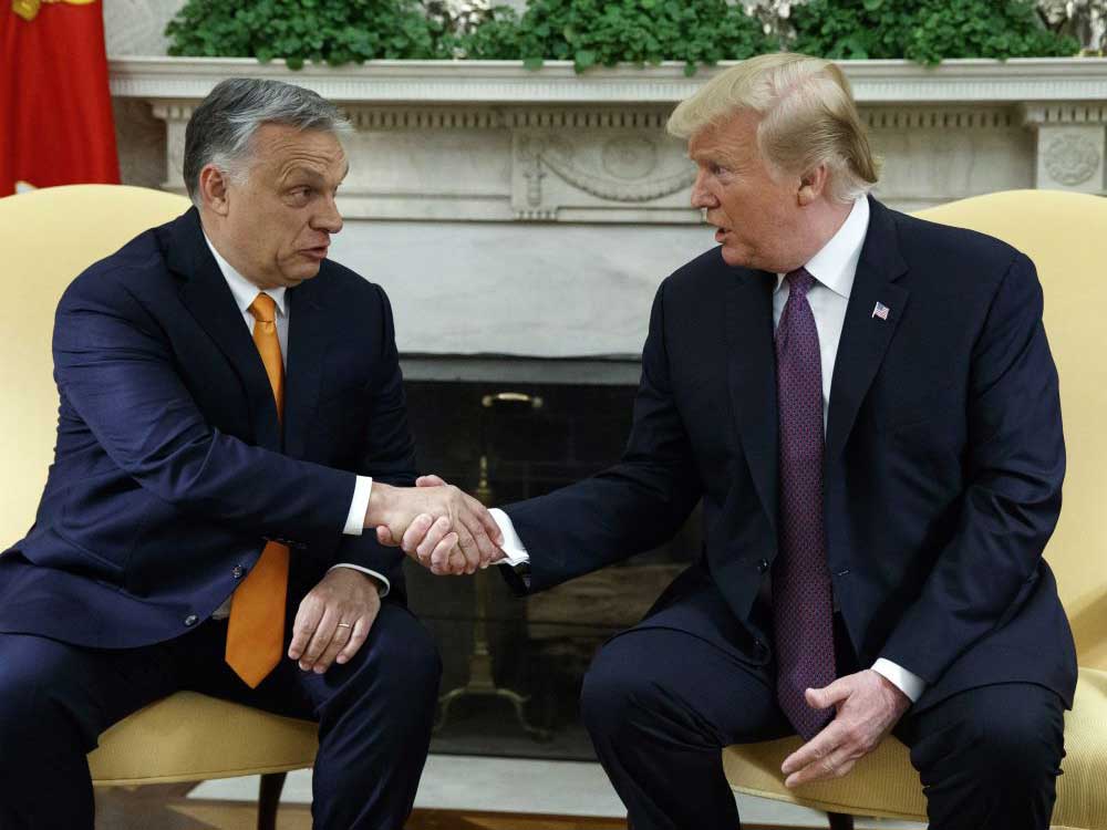 Two late-middle-aged white men in dark suits are seated and shake hands.