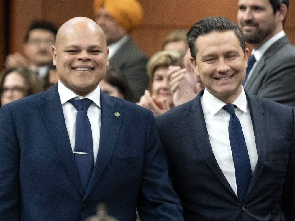 Two men, smiling, both in navy blue suits, with people applauding in the background. On the left, Jamil Jivani is bald with medium-light skin. On the right, Pierre Poilievre has light skin and short brown hair.