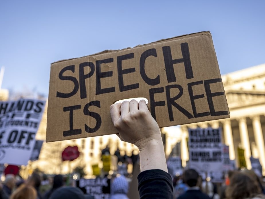 A person holds up a cardboard sign that reads &lsquo;Speech is free.&rsquo;
