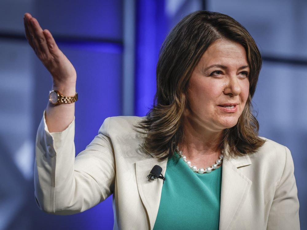 A light-skinned woman with shoulder-length brown hair, wearing an off-white blazer over a green blouse, gestures with one hand in the air.