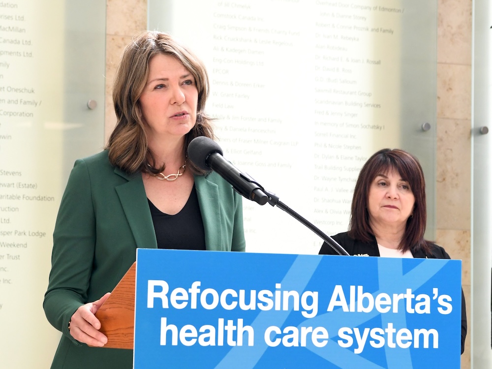 Danielle Smith, a mid-50s woman, stands at a podium with a blue sign saying ‘Refocusing Alberta’s Health Care System.’