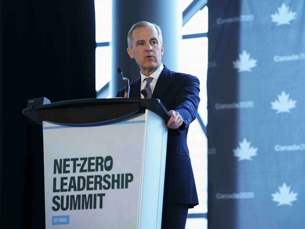 Mark Carney stands onstage at a white podium with a dark green sans-serif text that reads “Net-Zero Leadership Summit.” He is wearing a white shirt under a navy suit and tie. He has short grey hair and pale skin. Behind him is a projected blue digital wallpaper bearing the maple leaf reminiscent of the Canadian flag and the words “Canada2020” in sans-serif typeface.