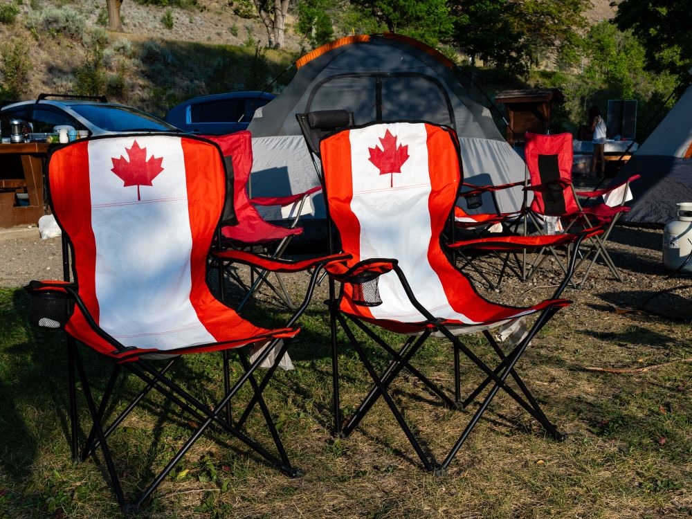 Two folding chairs bear patterns of the red-and-white Canadian flag. They are positioned next to each other on grass with a tent and parked cars behind them.
