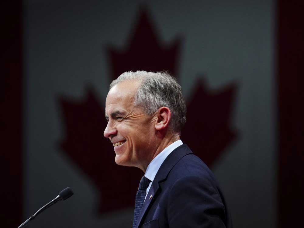 A side-profile shot of Prime Minister Mark Carney. Behind him, the silhouettes of a Canadian flag. 