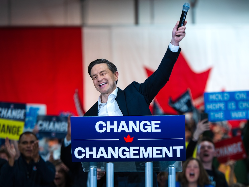 Pierre Poilievre, a smiling 45-year-old light-skinned man, holds a microphone far in the air as he stands behind a podium that says ‘Change’ and ‘Changement.’