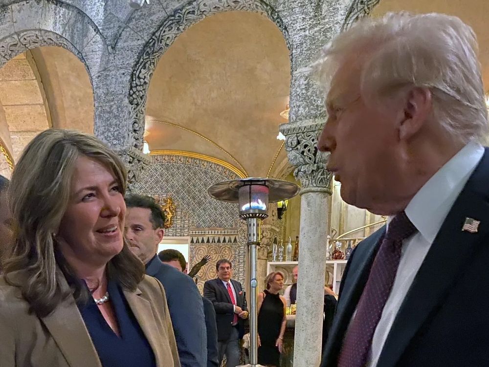 Danielle Smith stands with hands clasped and listens to Donald Trump in an ornate setting.