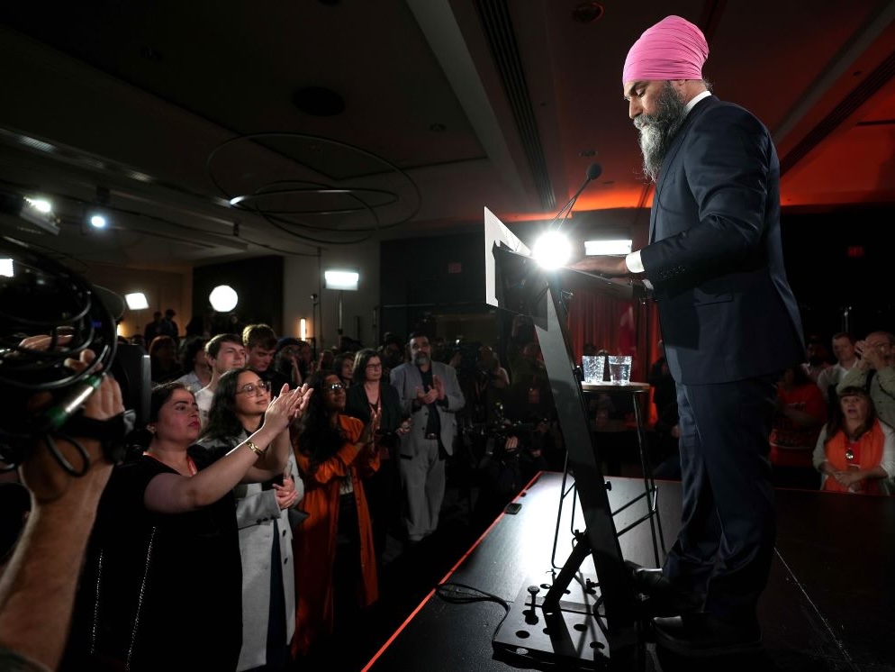 A sombre Jagmeet Singh stands at a lectern onstage, with supporters looking up from below.