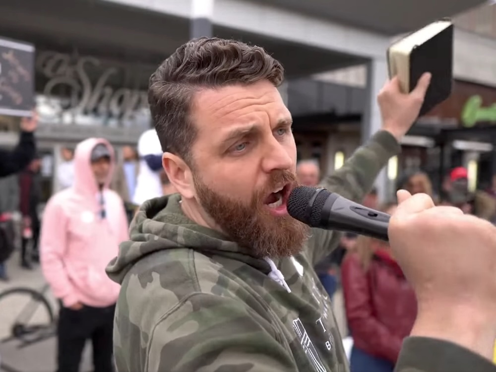 A 40-ish light-skinned man with trimmed hair and a beard, wearing a camo hoodie, speaks while holding up a Bible.