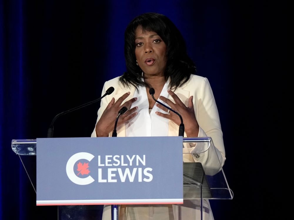 A woman in her 50s with dark skin and black hair wears a white suit and gestures as she speaks at a podium.