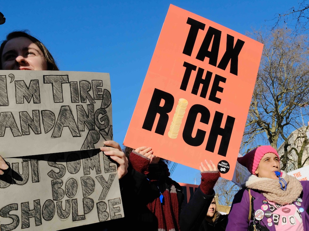 People are marching on a winter day. One holds an orange sign that says &lsquo;Tax the Rich.&rsquo;