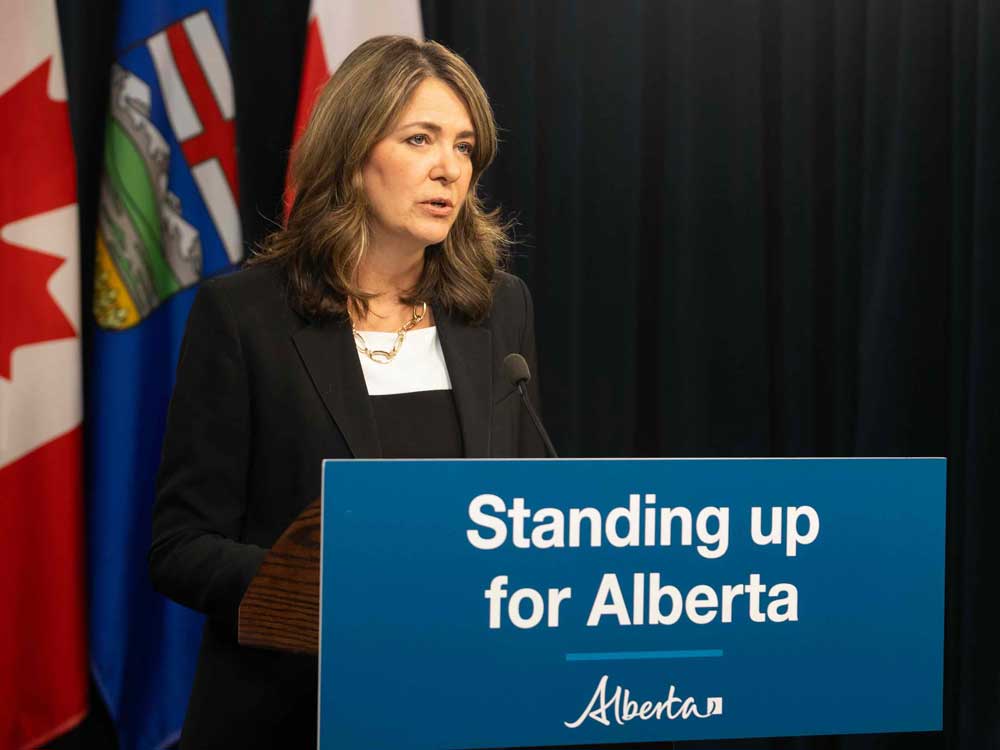 Danielle Smith has wavy, shoulder-length brown hair and a light skin tone. She is wearing a black blazer over a white top and standing behind a lectern on which a blue sign is affixed that reads &ldquo;Standing up for Alberta.&rdquo; Behind her are two Canada flags flanking one Alberta flag.