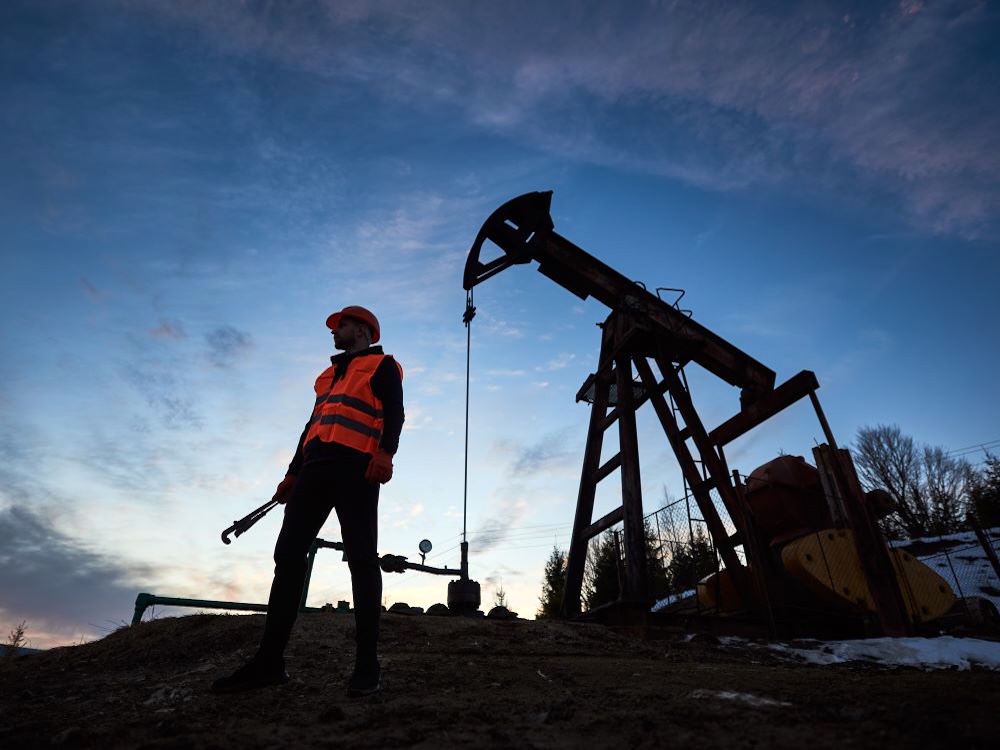 A person in a hard hat and high-vis vest, holding a large wrench, is silhouetted beside a grasshopper oil pump under a deep blue sky.