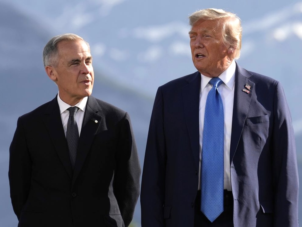 Mark Carney, a light-skinned man with short grey hair in his 60s, stands beside Donald Trump, a tall man with bronzed skin in his late 70s, with mountains in the background.