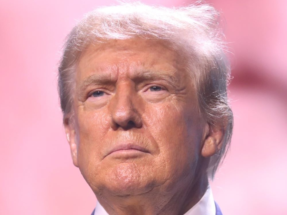 A close-up of the face of Donald Trump, a 79-year-old man with bronzed skin and platinum-silver hair.