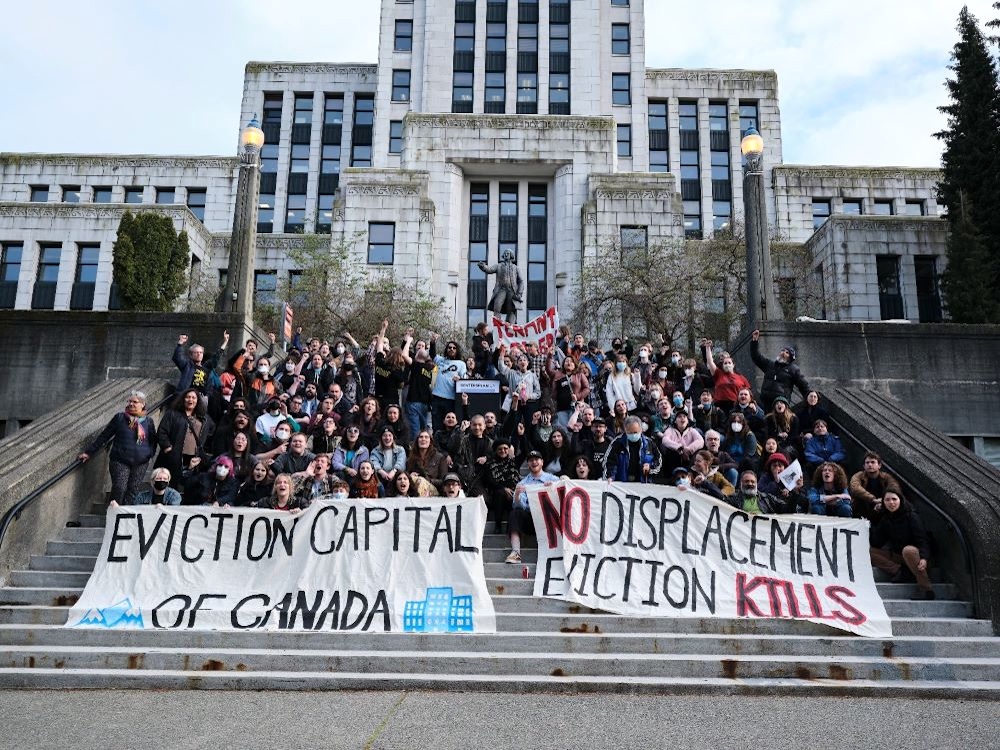 A group of people sit on the concrete steps in front Vancouver City Hall with large banners reading &lsquo;Eviction Capital of Canada&rsquo; and &lsquo;No Displacement. Eviction Kills.&rsquo;