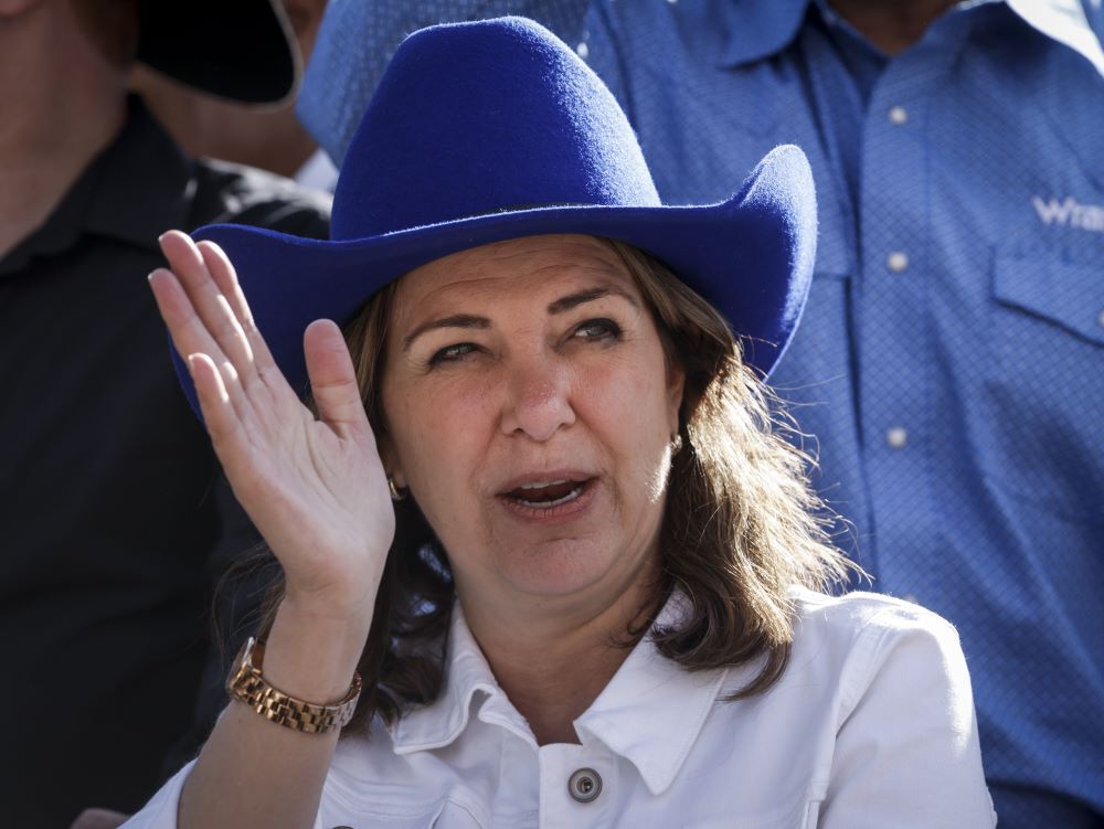 Medium close-up of Danielle Smith, a light-skinned 54-year-old woman with shoulder-length brown hair with blond streaks, wearing a blue cowboy hat and waving. 