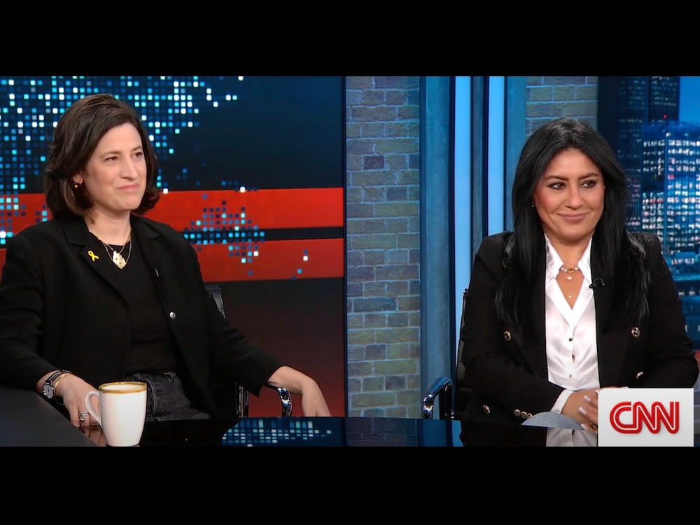 May Pundak and Rula Hardal are seated in a television studio against a brick wall and digital background featuring blue and red colourways and urban photography.