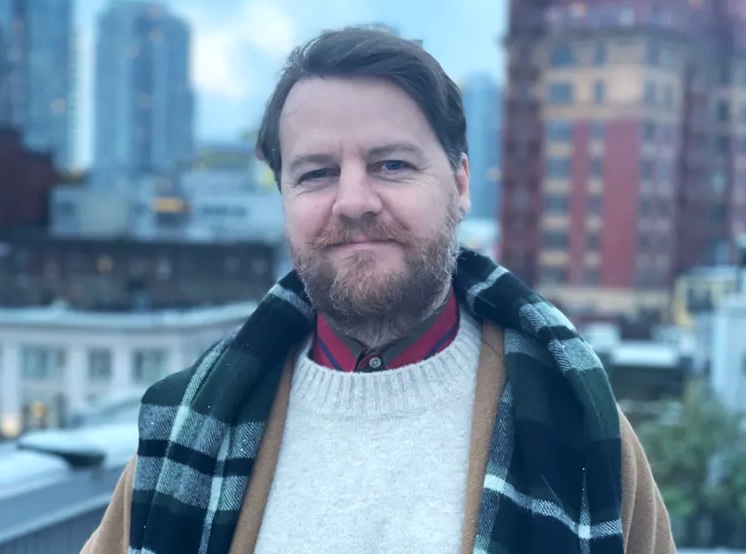 A 47-year-old light-skinned man with brown hair and a trimmed beard looks at the camera. He is wearing a jacket, a white sweater and a scarf draped over his shoulders.