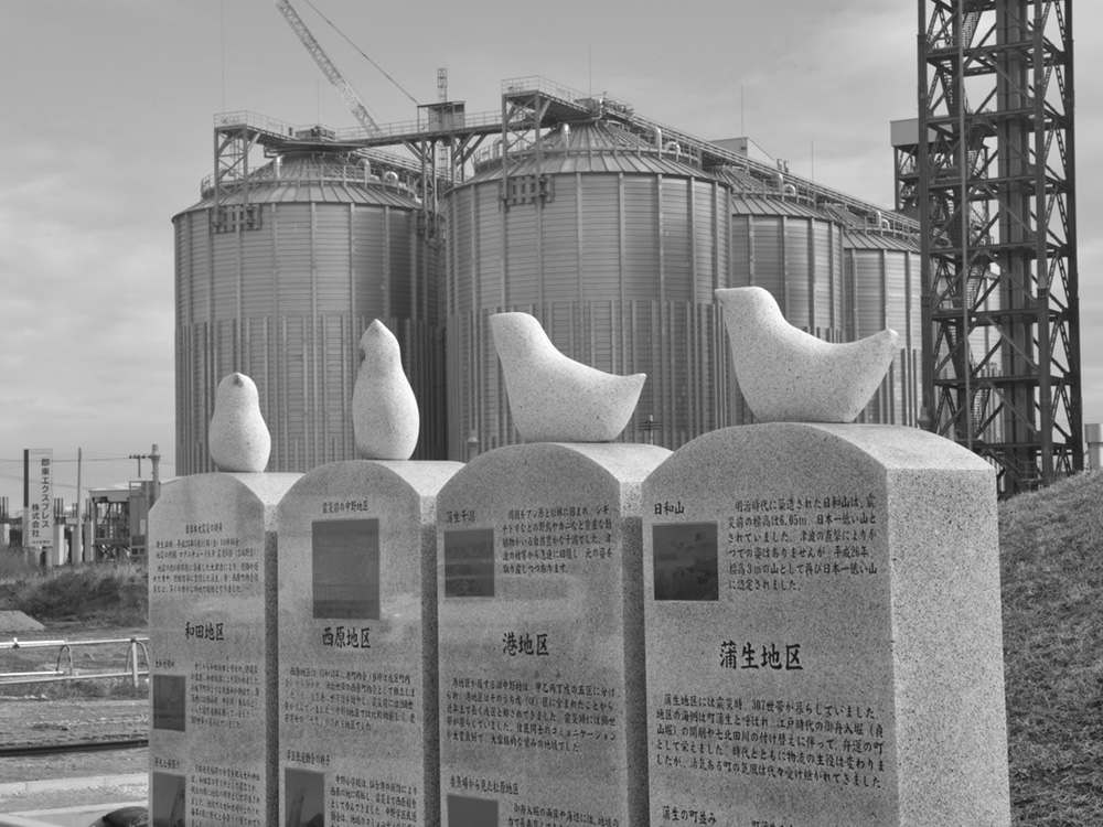A black and white photo shows four stone memorials topped with birds in the foreground. In the background, eight gleaming metal silos tower over the landscape.