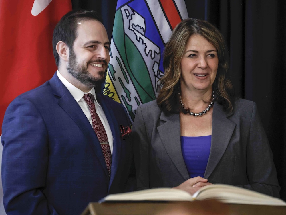 On the left, Alberta Education Minister Demetrios Nicolaides is standing in a navy suit over a white shirt and red tie, smiling. He has light skin tone, dark hair and a beard. He is standing at a lectern next to Alberta Premier Danielle Smith, who has medium-light skin tone, wavy brown hair and a grey blazer over a blue top.