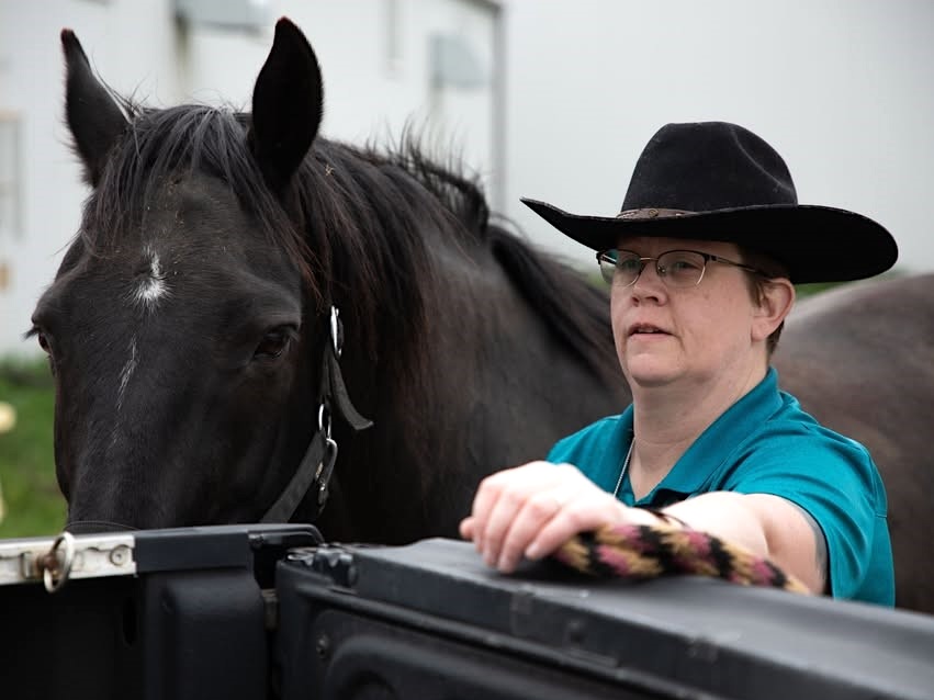A woman with light skin tone and short brown hair wearing a black cowboy hat and glasses stands next to a black horse.