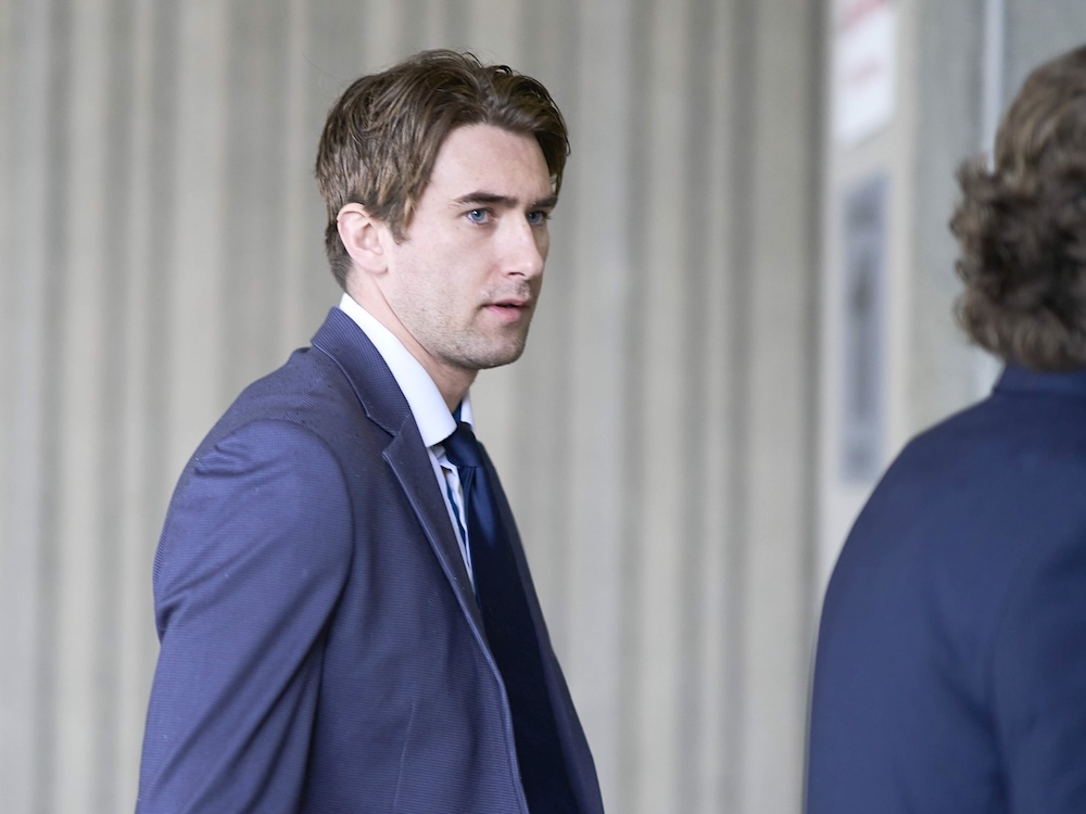 Michael McLeod is a man in his 20s with brown hair, a pale skin tone, blue eyes and a navy blue suit. He is walking with another person in a suit against a grey background.