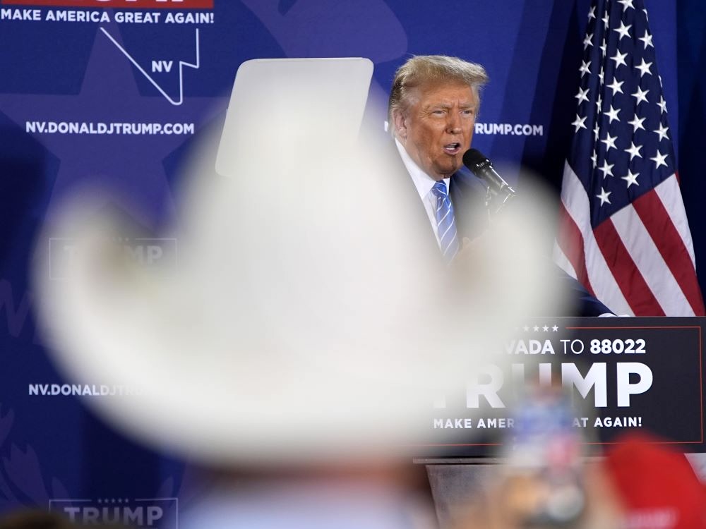 A view of Donald Trump speaking into a microphone at a lectern is partially blocked by a large out-of-focus white cowboy hat in the foreground.