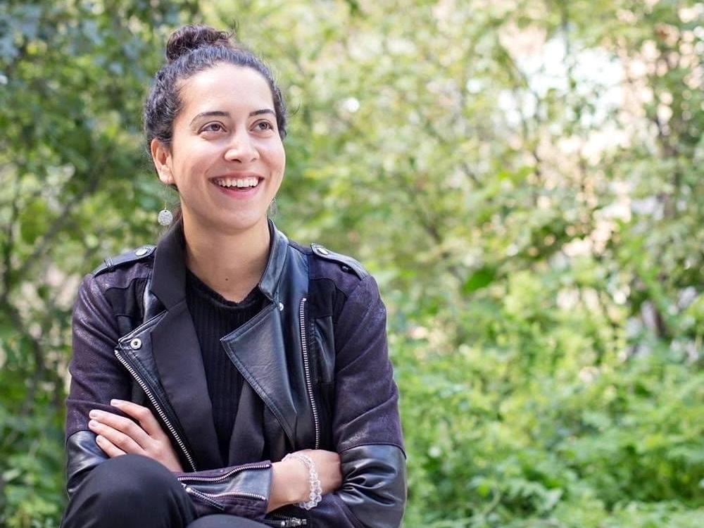 A dark-haired woman wearing a leather jacket and dark pants smiles while sitting in front of a tree-lined curb.