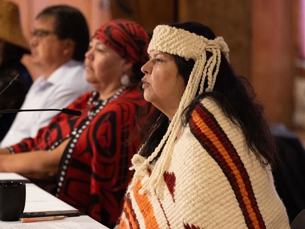 Indigenous leaders are seated next to each other at a table. One, wearing an Indigenous woven blanket and headband, speaks into a microphone.