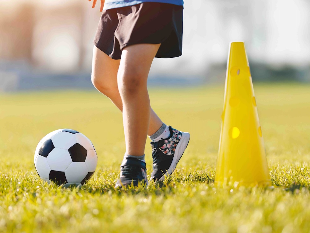 A child in shorts and black sneakers is visible from the waist down. They are standing between a yellow pylon in the grass and a black and white soccer ball.