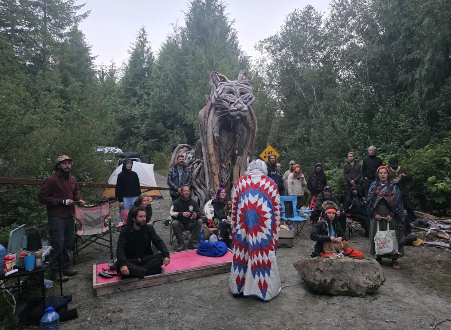 About 20 people stand around a giant wooden cougar sculpture, facing Bill Jones of the Pacheedaht First Nation, whose back is towards the camera.