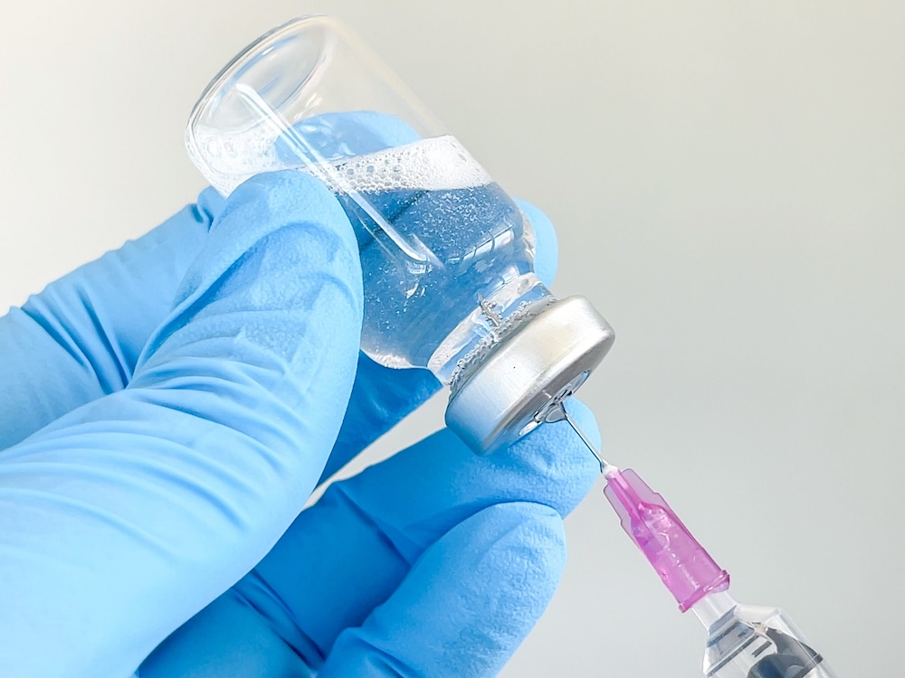 A hand wearing a blue medical glove holds a clear vial of fluid as it is tipped into the end of a medical syringe.