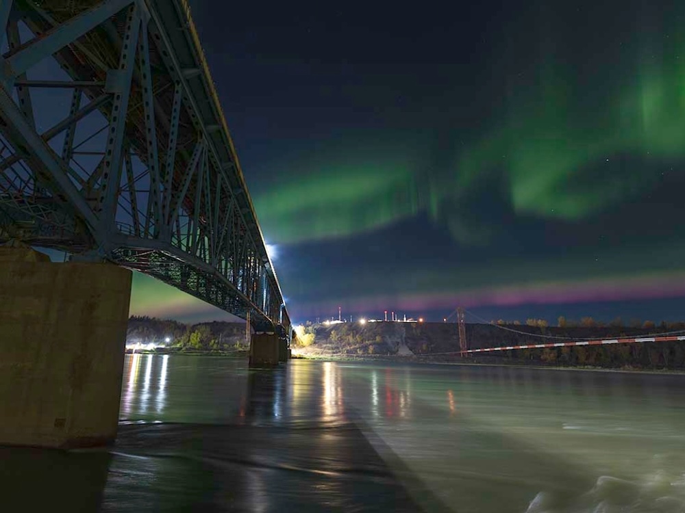 The Peace River flows at night under a bridge illuminated by the northern lights.