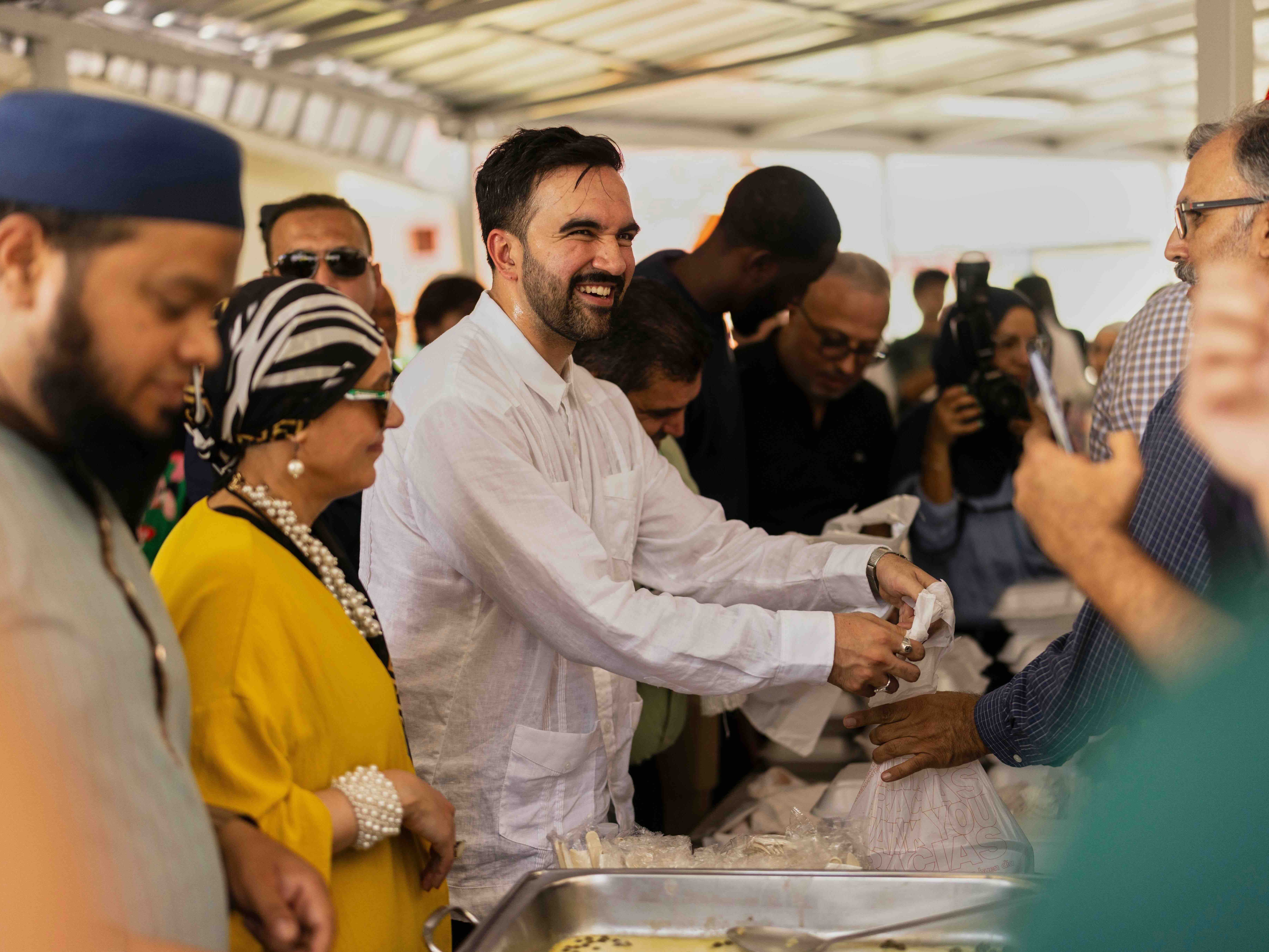 Zohran Mamdani is wearing a long white button-down shirt. He has short dark hair, a beard and a medium-light skin tone. He is standing inside a crowded white tent and handing a plastic bag containing food to a man to the right of the frame, standing on the other side of a table lined with food. Around Mamdani are people in cultural dress and headpieces.
