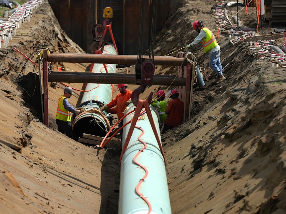 Workers in hard hats and high-vis jackets work alongside two lengths of pipeline inside a trench.