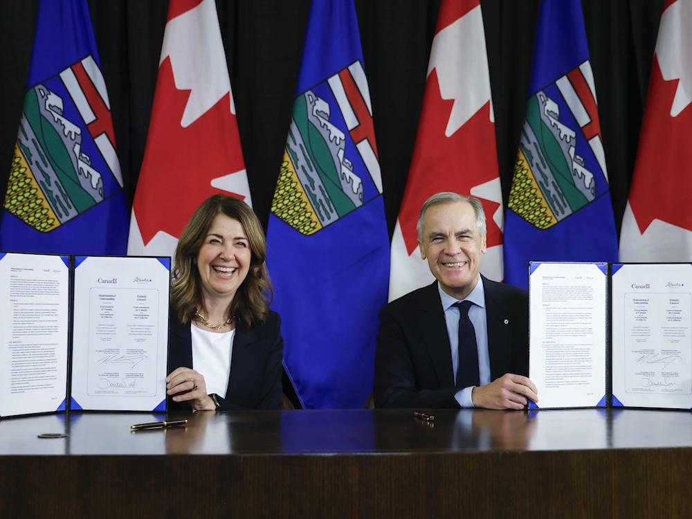 Danielle Smith, left, and Mark Carney, right, are seated at a dark wood table. They are each holding up a two-page document containing a memorandum of understanding that they have just signed. Smith has brown shoulder-length hair and a light skin tone. Carney has short grey hair with a light skin tone. They are smiling and seated in front of a row of Canadian and Albertan flags.