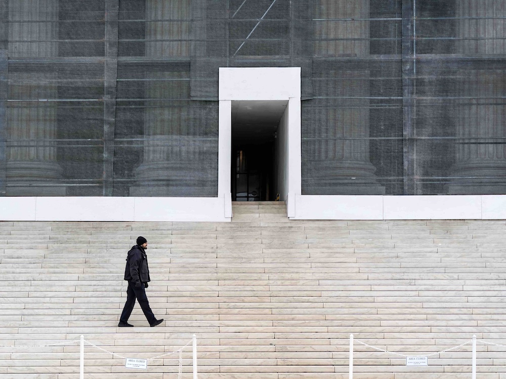 A police officer in a dark jacket and toque walks across a set of white stairs in front of a large white building with thick white columns. The columns are behind scaffolding and a dark grey construction screen.