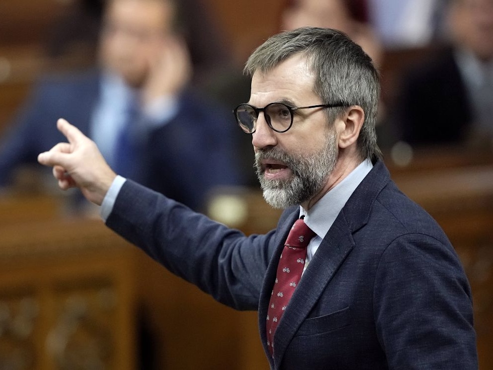 Steven Guilbeault, a man with light skin tone and salt and pepper short hair and beard, wearing glasses, suit and tie, points to his right while speaking.