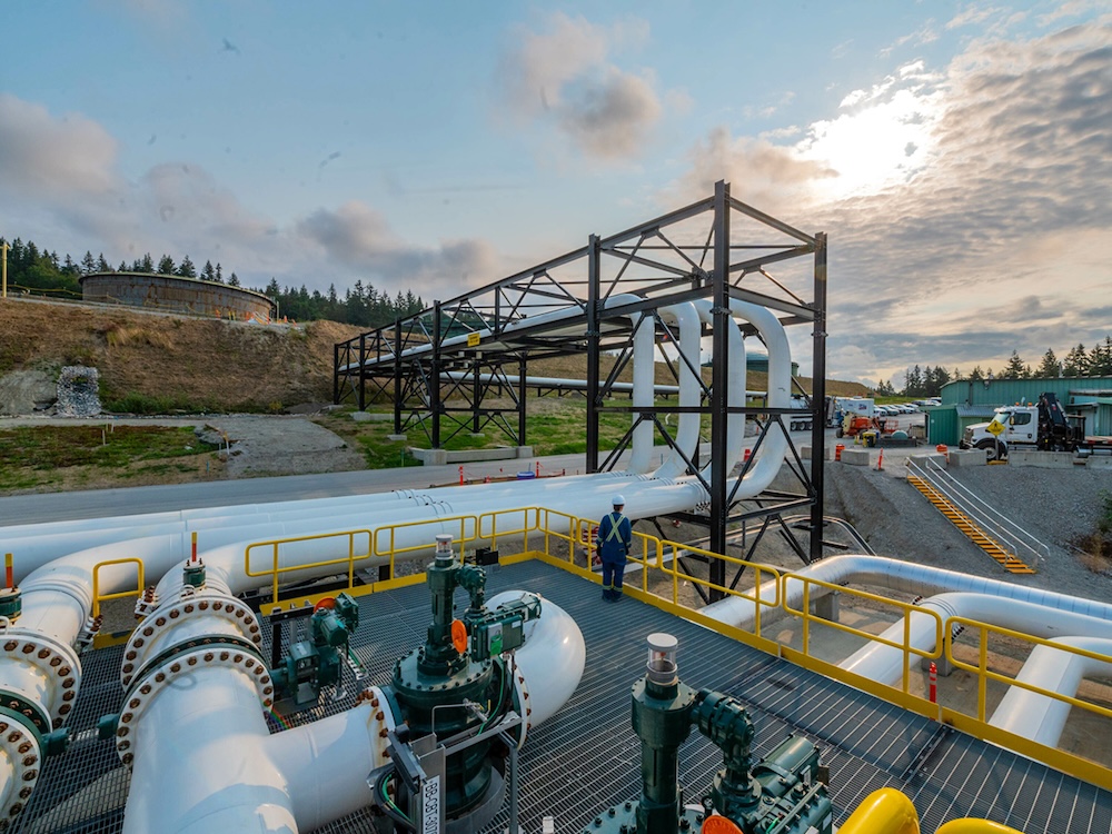 A facility with a tangle of large white pipes. In the background, a large tank on a hillside is visible.