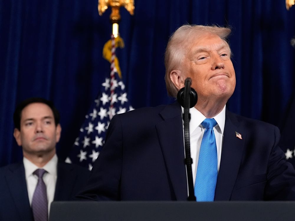 Donald Trump stands at a lectern, with lips pursed, an orange-tinged light skin tone and wispy white-blond hair. Marco Rubio looks on behind him, out of focus, with light skin and short dark hair.