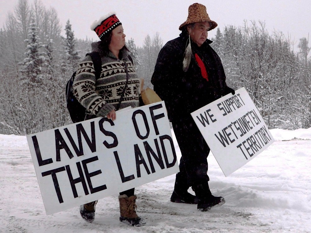 Two people hold signs and walk across a snowy landscape. The signs read &lsquo;Laws of the land&rsquo; and &lsquo;We support Wet&rsquo;suwet&rsquo;en territory.&rsquo;
