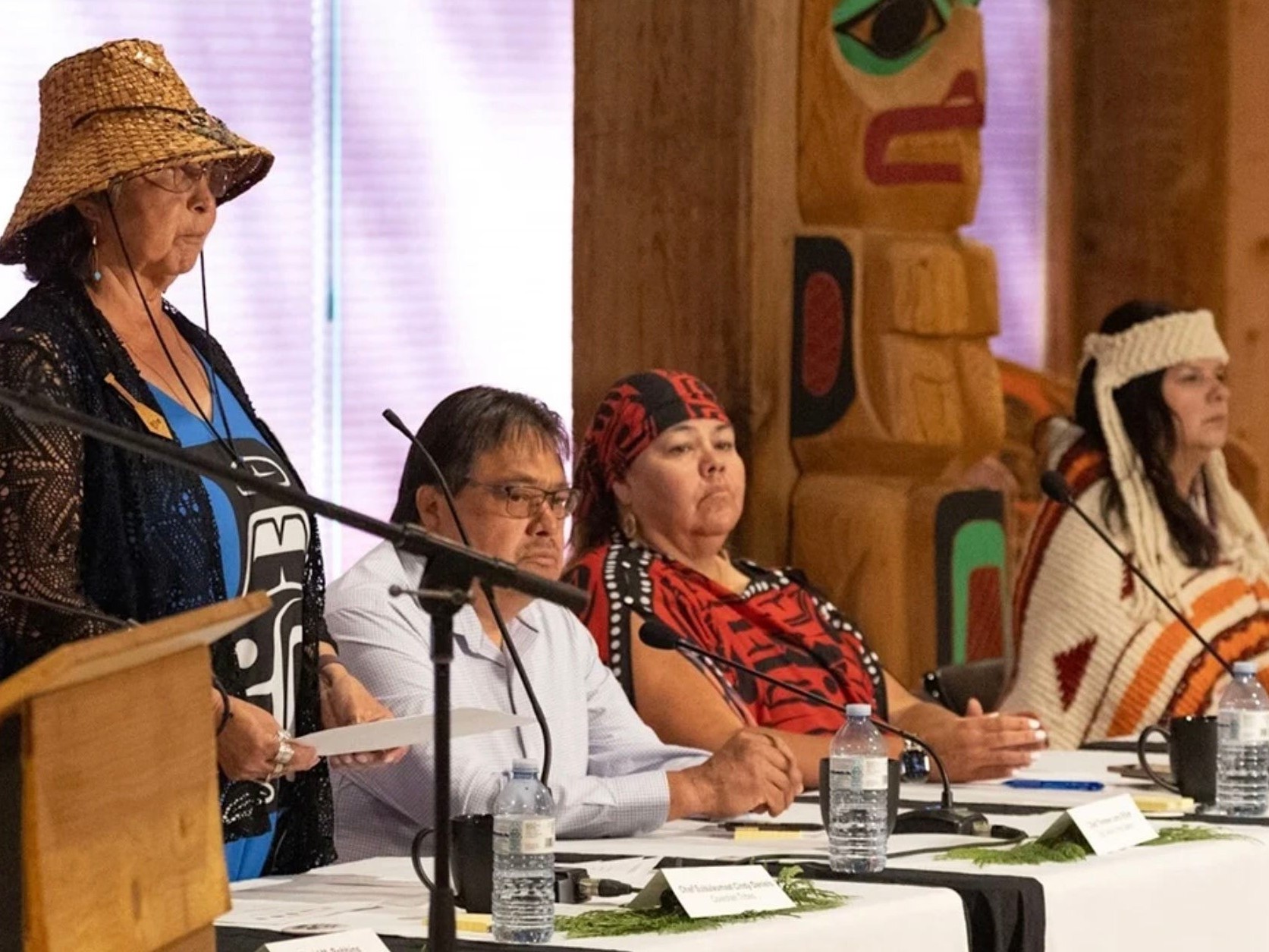 Four people are at a table in front of microphones. Three are sitting, one is standing to the left. 