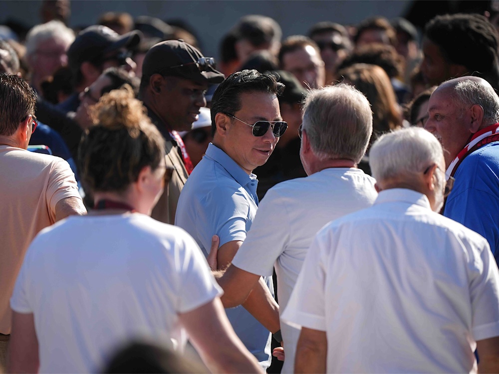 Ken Sim is wearing aviator sunglasses and a light blue short-sleeved button-down shirt. He is standing in a crowd of people with his back slightly turned to the camera. A man in a white T-shirt holds his arm.