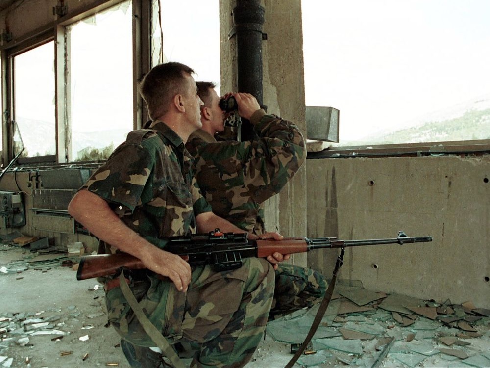 Two soldiers in fatigues crouch in a wrecked building by broken windows with glass shards all around them. One carries a rifle; the other looks through binoculars out a glass-less window.