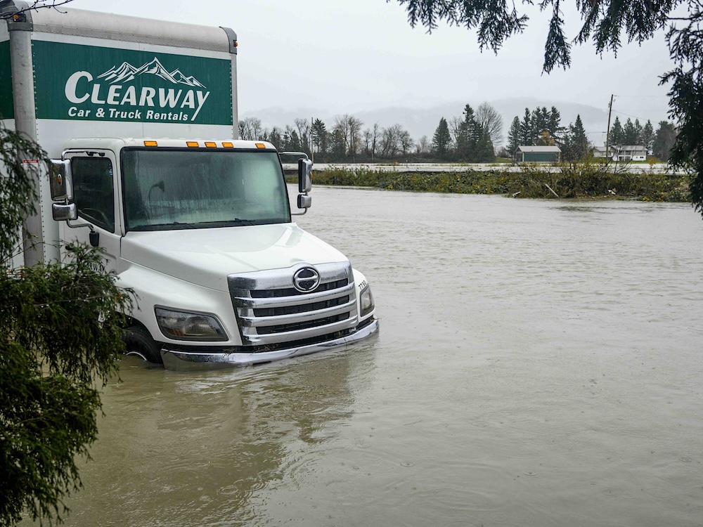 A large white Clearway rental truck stands submerged in grey water on a flooded piece of land. There are barns and houses in the background.