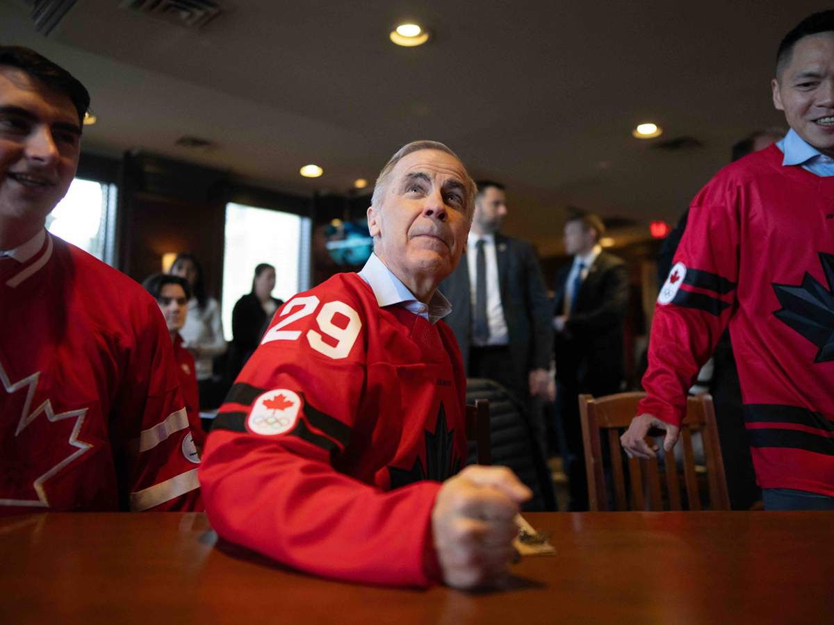 Mark Carney, centre, holds a fist on the table of a wooden bar inside a restaurant. He is wearing a red Team Canada hockey jersey over a light collared shirt.