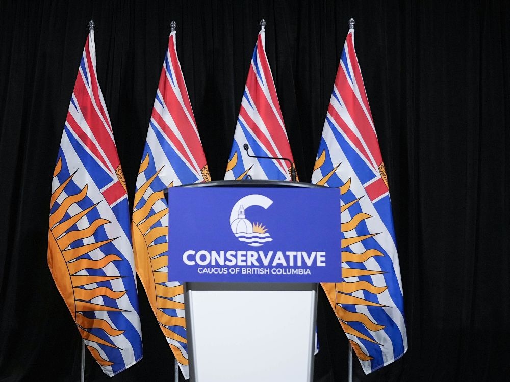 A podium with a sign saying &lsquo;Conservative caucus of BC&rsquo; stands in front of four B.C. flags. No one is behind it.