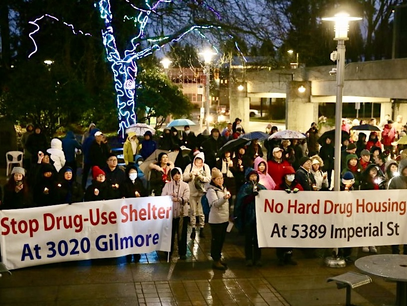 Protesters hold signs that read &lsquo;Stop drug-use shelter at 3020 Gilmore&rsquo; and &lsquo;No hard drug housing at 5389 Imperial St.&rsquo; It&rsquo;s a winter night and many are wearing rain jackets or holding umbrellas.