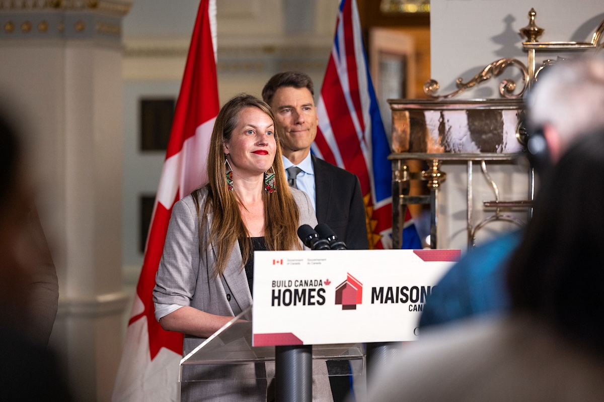 Christine Boyle stands at a podium with a &lsquo;Build Canada Homes&rsquo; sign on it. She is wearing a grey suit and has long blond hair. Behind her Gregor Roberston stands facing the audience. He is wearing a navy suit.